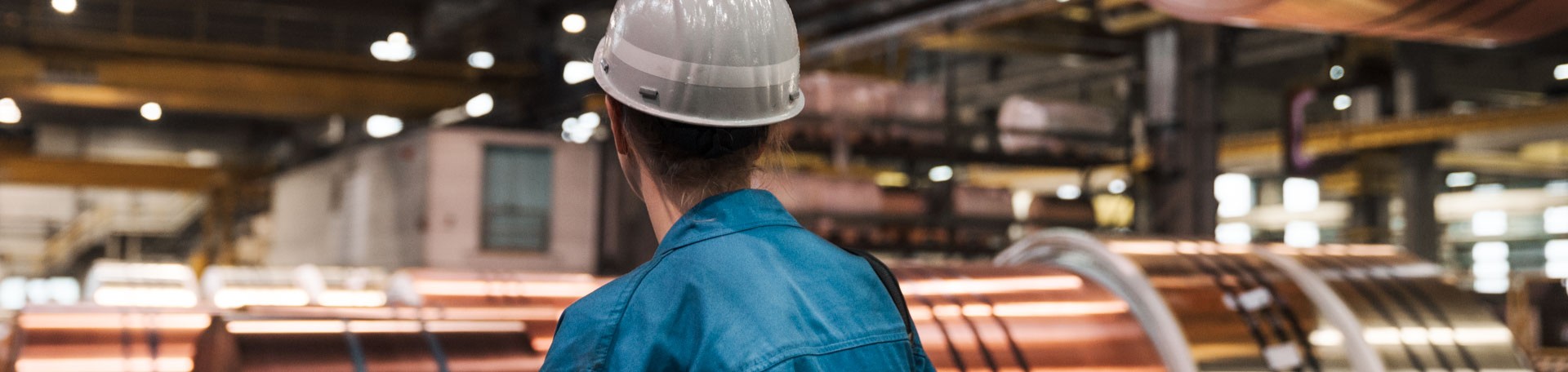 Arbeiterin mit Schutzhelm in Halle, umgeben von großen Kupfercoils.