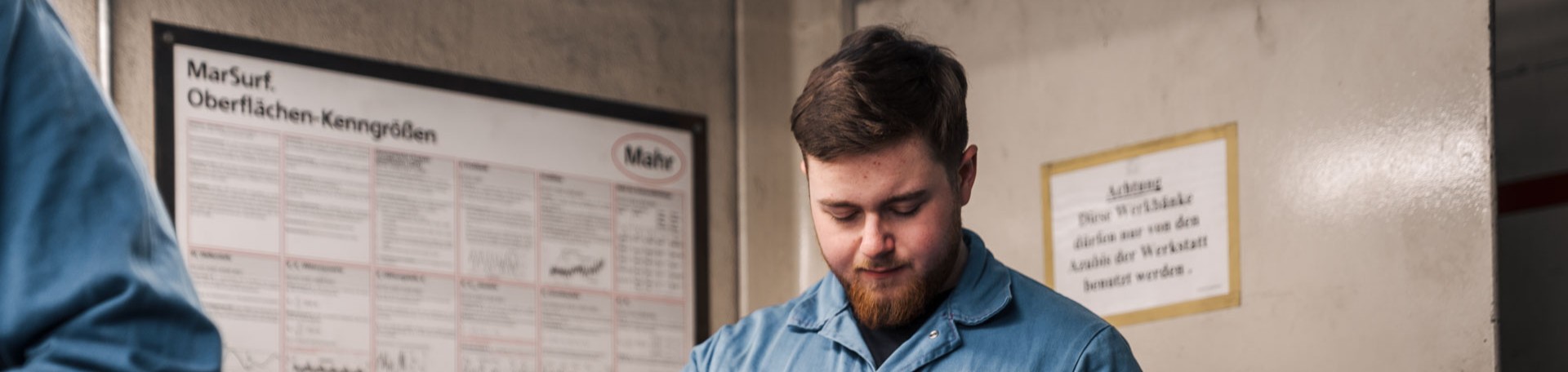 Azubi in blauer Arbeitskleidung arbeitet konzentriert vor einer Wand mit technischen Schildern.