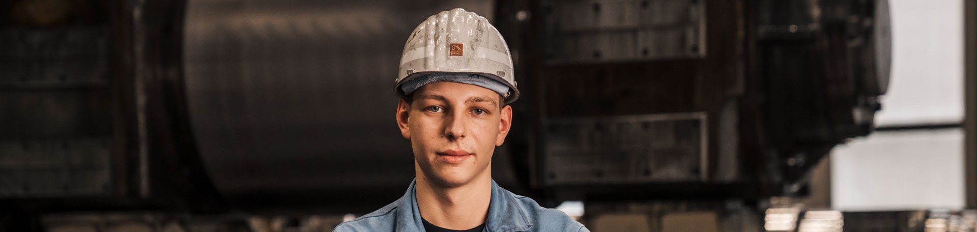 Arbeiter mit Schutzhelm steht in einer Fabrikhalle vor industriellen Maschinen.