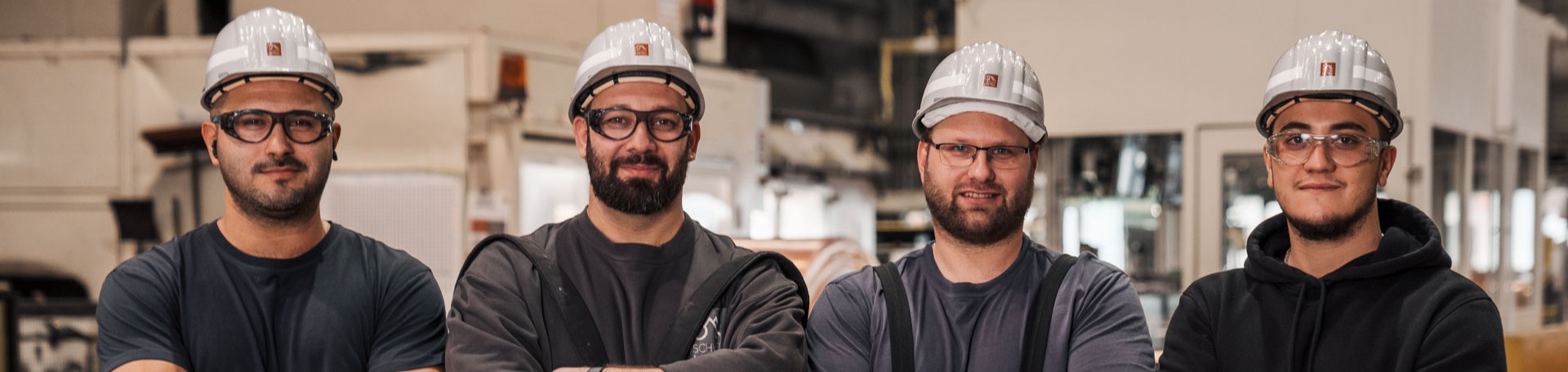 Vier Arbeiter mit Schutzhelmen und Brillen lächeln in einer Fabrikhalle in die Kamera.