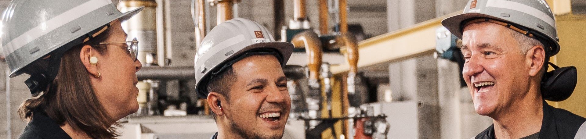 Drei Personen mit Schutzhelmen und Ohrstöpseln lachen in industrieller Umgebung.