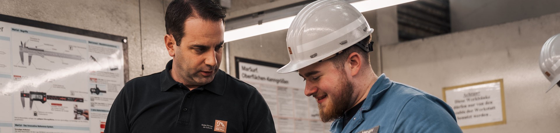 Mitarbeiter erklärt Azubi mit Schutzhelm etwas in Werkstatt vor technischen Postern.